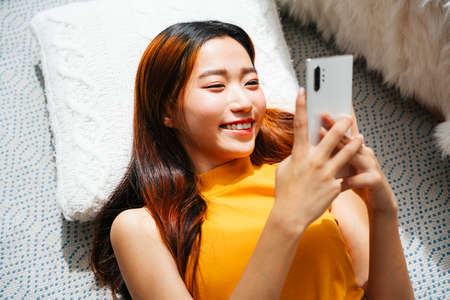 Beautiful young asian woman laying on a pillow using smartphone at home.の写真素材