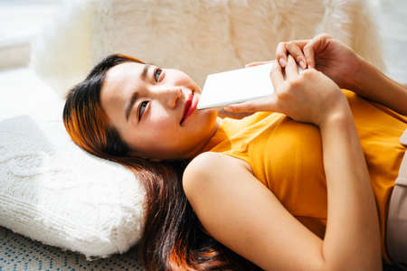 Beautiful young asian woman laying on pillow holding smartphone.の写真素材