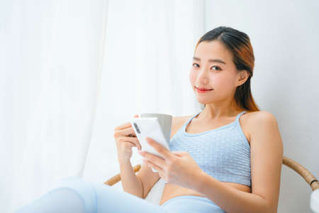 Beautiful sporty asian woman wearing sportbra holding smartphone and coffee cup.
