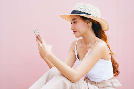 Side view - Beautiful asian woman wearing hat using smartphone isolate on pink background.の写真素材