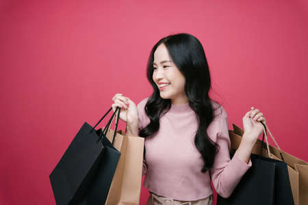 Asian shopping lover enjoy buying stuff in mall. Shopaholic girl holding many paper bags isolate on pink background.の写真素材