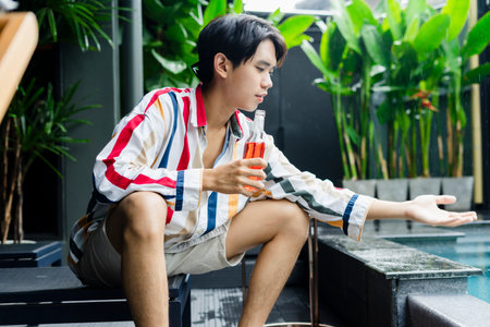 Cheerful asian young man enjoy bottle of wine, sitting near swimming pool at home.の写真素材
