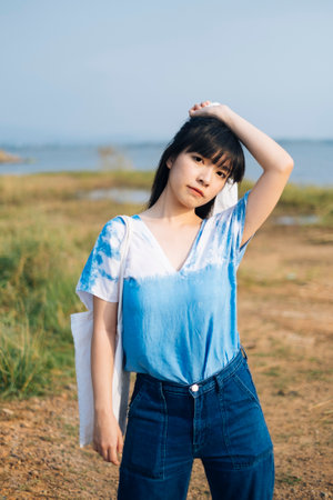 Black half tied hair girl in white and blue tie dye standing in grassland next to the sea and raise canvas bag at her head.の写真素材