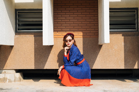 Wavy brunette woman in blue and red retro dress sitting in the sunlight next to the brick building.の写真素材