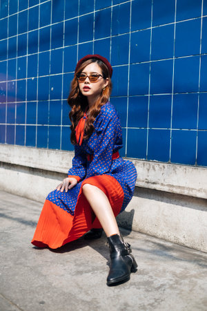 Wavy brunette woman in blue and red retro dress and beret sitting at the blue wall in the sunlight with sexy posture.の写真素材