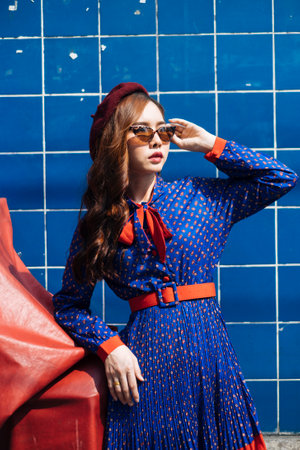 Wavy brunette woman in blue and red retro dress and beret standing at the blue wall in the sunlight prop her elbow on red box.の写真素材