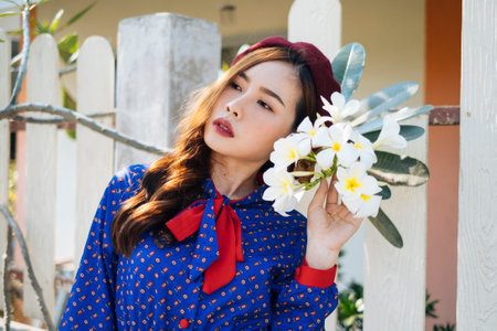 Wavy brunette woman in blue and red retro dress standing at the white fence and snuggling up to plumeria.の写真素材
