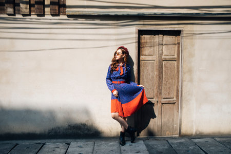 Wavy brunette woman in blue and red retro dress standing on the slope path in front of wooden dooe.の写真素材