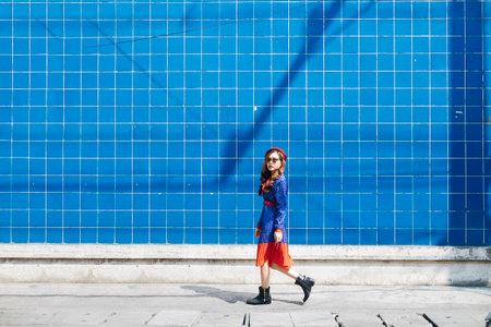 Wavy brunette woman in blue and red retro dress and beret walk along the blue wall in the sunlight.の写真素材