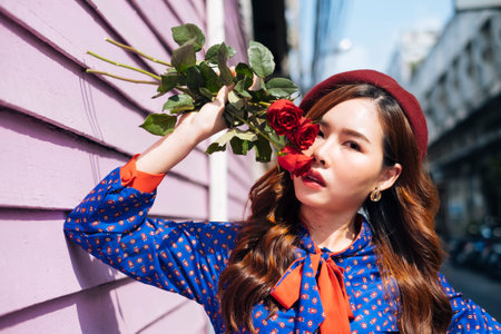 Wavy brunette woman in blue and red retro dress hold red roses at her face and standing at the pink...
