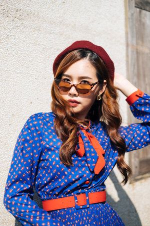 Medium portrait shot of wavy brunette woman in blue and red retro dress standing with cool posture at the old house's wall in sunlight.の写真素材