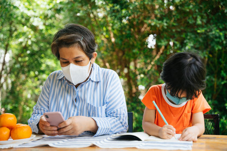Senior elderly old woman wear mask using smartphone and grandson drawing something on paper at yard.の写真素材