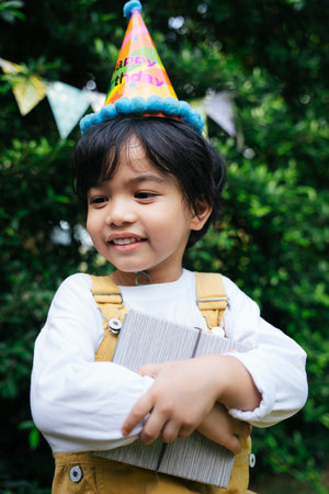 Cheerful asian black hair kid wear party hat holding a gift box at yard.