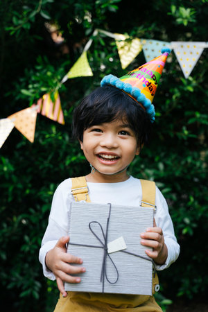 Asian black hair kid wear party hat holding a birthday gift box at yard.の写真素材