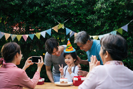 Asian kid celebrate birthday party with family outdoors. Mother taking picture with smartphone.の写真素材