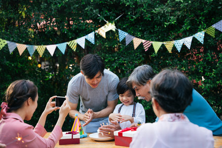 Asian kid celebrate birthday party with family outdoors. Mother taking picture with smartphone.