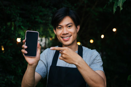 Cheerful asian waiter wear black apron showing blank screen mock up smartphone for copy spaceの写真素材