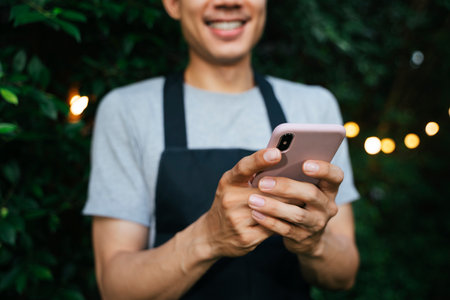 Portrait of cheerful smile asian waiter wear black apron usign smartphone at yard restaurant.の写真素材
