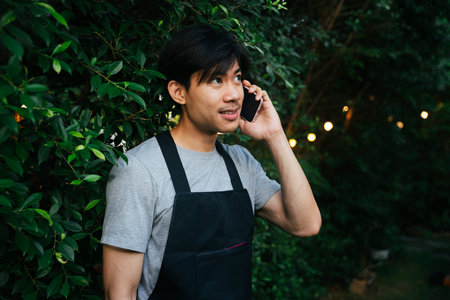 Cheerful asian waiter wear black apron talking on the phone at yard outdoors.の写真素材