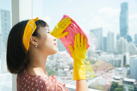 Maid service woman wearing yellow protection gloves cleaning glass window with dry rag in apartment.の写真素材