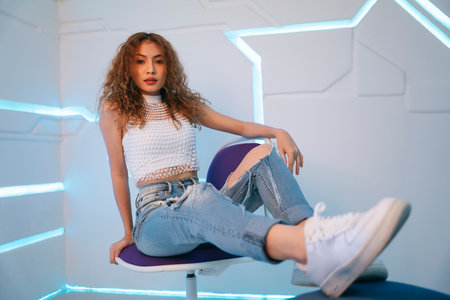 Fashion portrait of young asian curly hair woman with jeans pant sitting on a chair.の写真素材