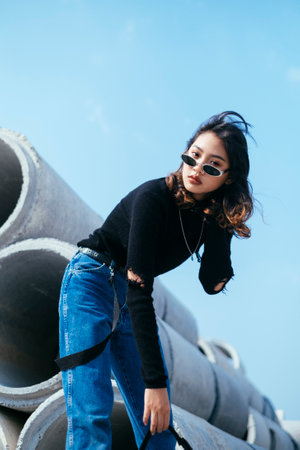 Low angle shot of black hair girl in sweater and jeans standing and bend down her body on the big concrete tube.の写真素材