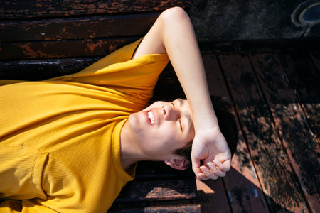 Guy in yellow t-shirt and blue pants lie down on the wooden bench in sunlight, raise his arm at forehead.の写真素材