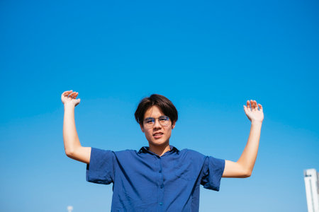 Black hair guy in blue clothes and eyeglasses raise his hands to the blue sky.の写真素材