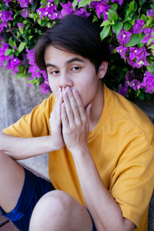 Guy in yellow t-shirt and blue pants sitting on the wooden floor leaning against the barricade of pink bougainvillea, close his mouth.の写真素材