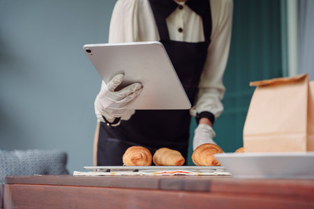 Bakery employee woman checking order on digital laptop for online delivery. Fresh baked croissants.の写真素材