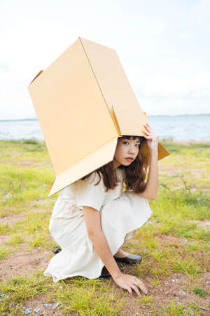 Young asian thai woman hipster curly hairstyle on white dress putting cardboard box over head. Sitting posing. Cute girl at lake.の写真素材