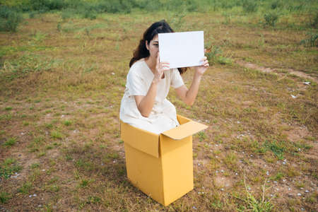 Young asian thai woman hipster curly hairstyle on white dress sitting inside the box showing blank paper sign. Copy space.の写真素材
