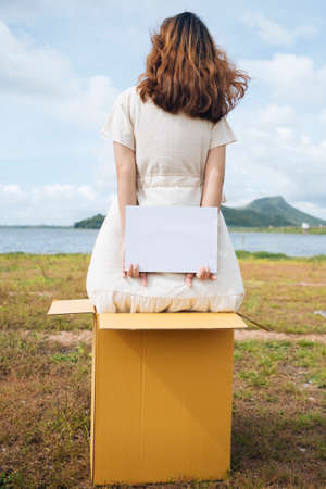 Back view - Young asian thai woman hipster curly hairstyle on white dress holding blank paper sign behind the box at lake. Copy space.の写真素材