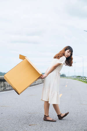 Side view of Young asian thai woman hipster curly hairstyle on white dress carrying cardboard box behind the back.の写真素材
