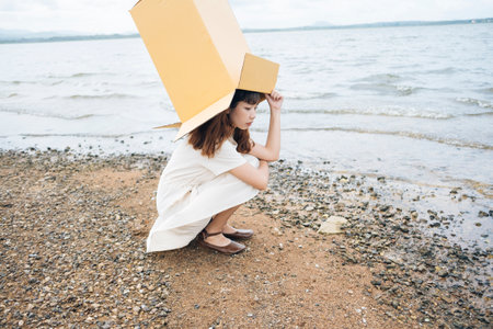 Side view of Young asian thai woman hipster curly hairstyle on white dress putting cardboard box over head sitting at lake.の写真素材