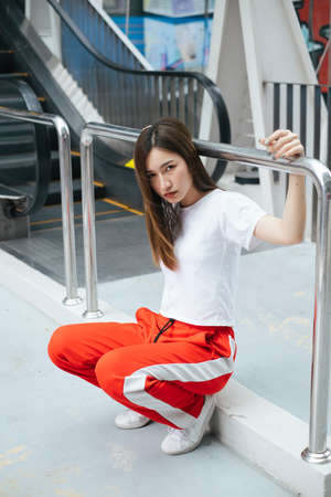 Fashion portrait - Beautiful asian young woman in white shirt and red sport pant sitting on overpass in the city.の写真素材