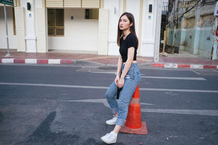 Full length fashion portrait - Beautiful hipster asian young woman in black shirt and jeans pants sitting on red traffic cone on the road.の写真素材
