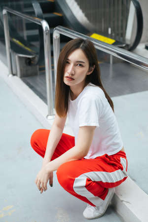 Fashion portrait - Beautiful asian young woman in white shirt and red sport pant sitting on overpass in the city.の写真素材