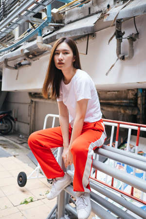 Full length fashion portrait - Beautiful asian young woman in white shirt and red sport pant sitting on rail bar at street alley.の写真素材