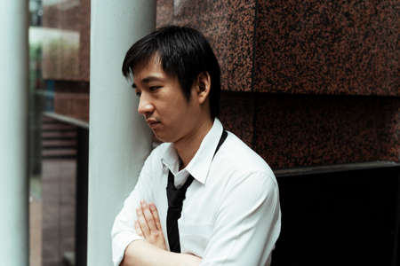 Stressful business man standing next to white pole on the street. Asian business man on white t-shirt facing the pole. Sadness man feeling depress wandering the street.の写真素材