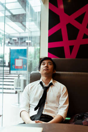 Thai local business man sitting in an modern office looking through the glass window. Asian business person having burn out situation. Business man feeling exhausted. Thai business man looking depress.の写真素材