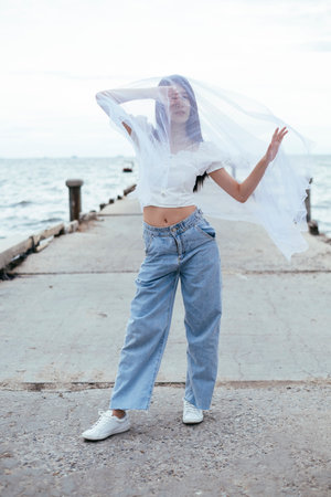 Portrait of young asian long dark hair woman with white veil at sea bridge. Shirt and blue jeans...
