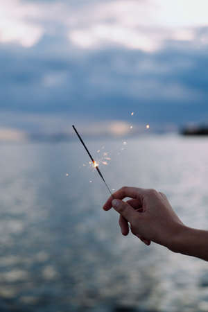 Hand holding a sparkler with blurred deep sea ocean water background at dusk time.の写真素材
