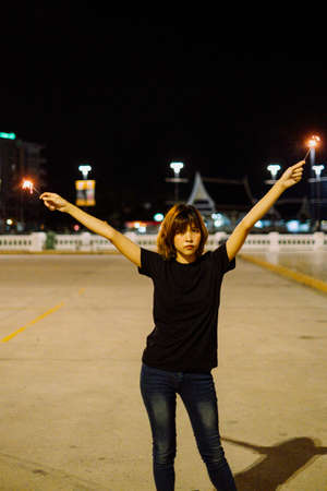 Young asian woman short hair wearing black shirt and jeans pants holding two sparklers raise up hands at night.の写真素材