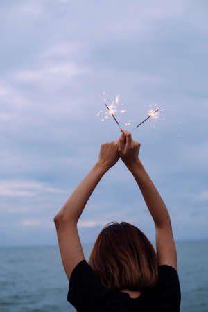 Back view of young asian thai woman short hair raise hands holding two sparklers over head on the beach at dush time.の写真素材