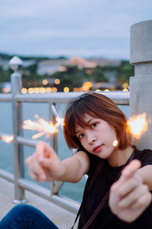 Young asian thai woman short hair sitting on sea bridge playing with sparklers at dusk time.の写真素材