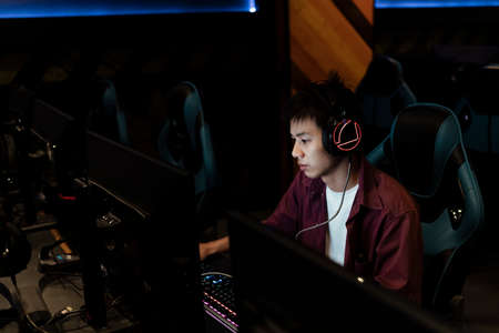 Young male asian gamer playing computer at internet cafe with serious looking, wearing headphone, sitting on gamer chair.の写真素材