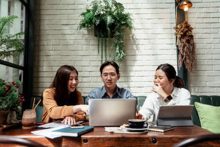 Front view of three young business people working together, looking on laptop. Meeting taking place in cafe. Three young business people discussing about business idea and concept on wooden table.の写真素材