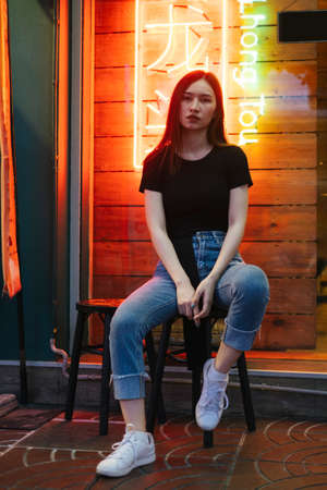 Fashion portrait - Beautiful hipster asian young woman in black shirt and jeans pants sitting on chair over neon light background.の写真素材