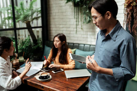 Three young asian business people using tablet in the meeting to explain their business concept and idea. The meeting taking place in cafe. Young business boy making presentation to their colleagues.の写真素材